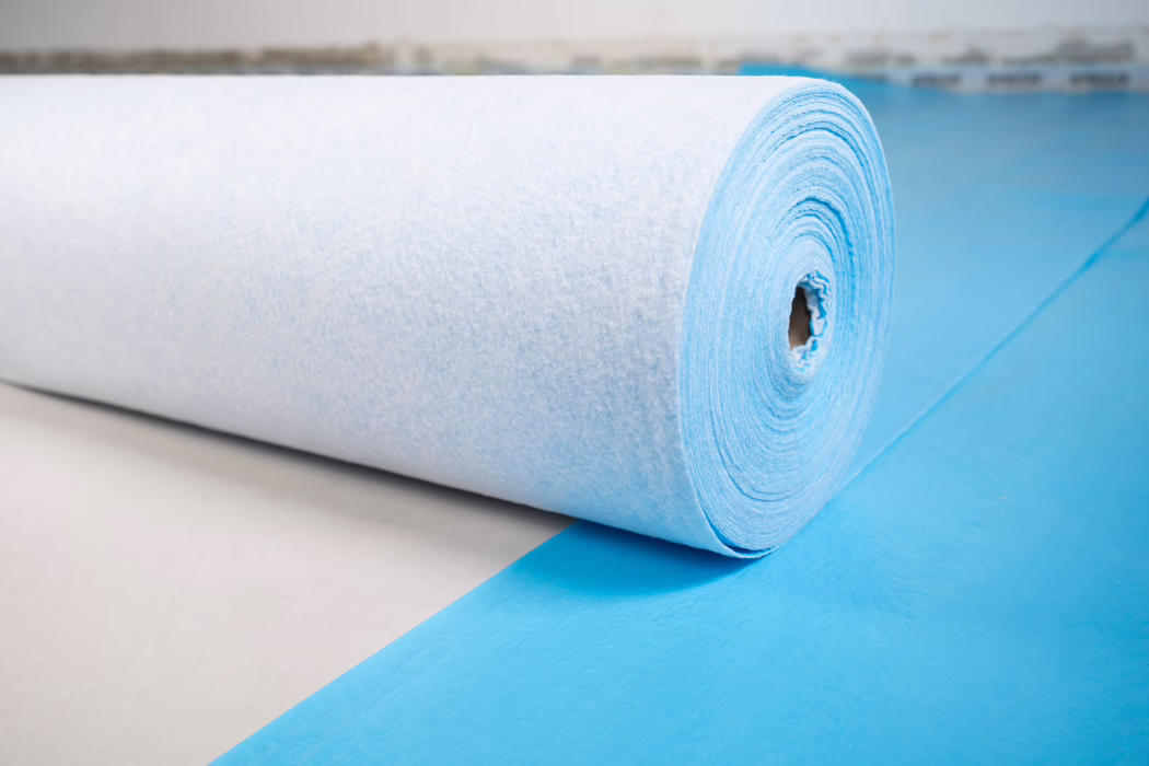 Detailopname van een grote rol lichtblauw non-woven stof of onderlegmateriaal op een bijpassende blauwe ondergrond. De foto toont het gestructureerde, vezelige oppervlak en de spiraalvormige wikkelingen rond een papieren kern, geplaatst op een lichte vloer. Zacht, diffuus licht benadrukt de fijne vezels en de cilindrische vorm; de achtergrond is onscherp.
