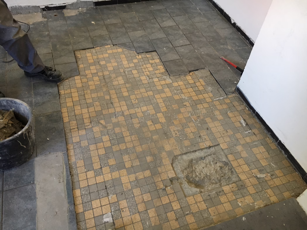 UZIN_Application_Image_UZIN NC 110_0445_ 2018-03 Een gedeeltelijk verwijderde tegelvloer tijdens renovatiewerk: op de voorgrond kleine beige en grijze mozaïektegels met versleten en ontbrekende delen; rondom liggen grotere donkere rechthoekige tegels los, waardoor lijm en ondervloer zichtbaar zijn. Links zijn de benen en zwarte werklaarzen van een medewerker zichtbaar naast een emmer met verwijderde tegels; rechts leunt een rode koevoet tegen de muur. Witte muren met zwarte plint, stof en puin verspreid over de vloer. Foto genomen vanuit een laag perspectief naar beneden.