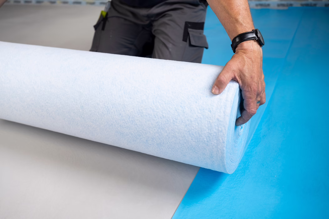 Close-up van de hand van een arbeider die een grote lichtblauw-witte onderlaag- of isolatierol afrolt op een felblauwe vloer. De rechterhand draagt een zwarte polshorloge en een eenvoudige ring en houdt de rand van de rol vast; een grijze werkbroek met zakken en een vastgeklikt gereedschap zijn deels zichtbaar op de achtergrond. De zachte, vezelige structuur van het materiaal is zichtbaar, de scène doet denken aan binnenwerkzaamheden of vloeren leggen en de compositie benadrukt de kromming van de rol en het kleurcontrast tussen wit, lichtblauw en felblauw van de vloer.