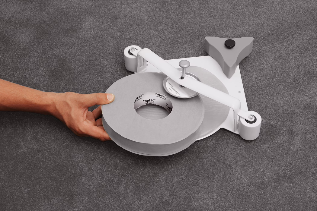 UZIN_Application_Image_Toptac 35_2_2018-03 Close-up van een linkerhand die een grote grijze rol plakband vasthoudt, geplaatst op een laag profiel tape-dispenser op een grijs, gestructureerd stoffen oppervlak. De dispenser heeft een wit metalen arm met een centrale spil en een rond snijwiel, twee kleine witte rolletjes aan de basis en een driehoekige grijze stabilisator met een zwarte rubberen voet; de kartonnen kern van de tape toont het merk 'Toptac'. Zacht, gelijkmatig licht benadrukt het neutrale palet van grijzen en wit. De compositie richt zich op de rol en de hand linksonder, met zichtbare huidtint en aderen zonder identificeerbare kenmerken.