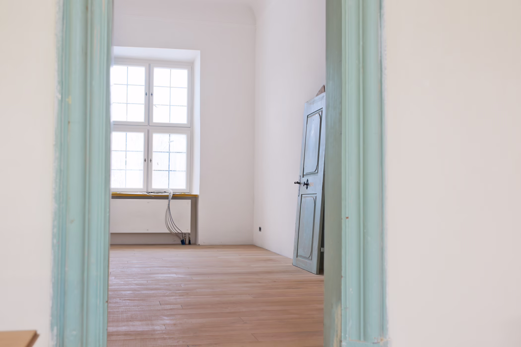 UZIN_Application_Image_MK 200_02_2018-10 Interieurzicht door een licht turquoise geschilderde deurpost naar een lege, zonovergoten kamer. Een groot vierdelig raam in het midden van de achterwand vult de ruimte met zacht natuurlijk licht. De kamer heeft licht geschuurde houten vloerplanken, witte pleistermuren en een licht verweerde blauwgrijze houten deur die tegen de rechterwand leunt. Bij het raam liggen lage verwarmingsbuizen en een bundel blootliggende elektrische kabels op de vensterbank en lopen naar de vloer, wat duidt op lopende renovatiewerkzaamheden. De algehele sfeer is rustig en minimalistisch, met gedempte pasteltinten en strakke architectonische lijnen.