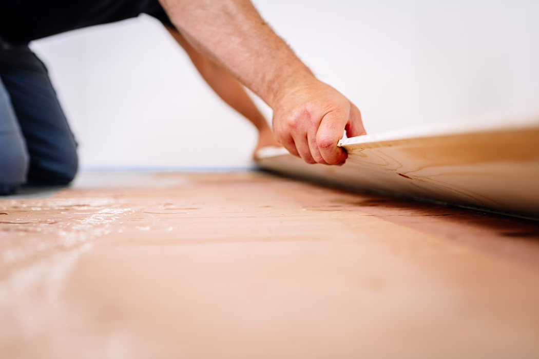 UZIN_Application_Image_MK_250_11_2021-11 Nabijbeeld van iemand die vloer legt: de hand van een knielende vakman grijpt de rand van een laminaat- of houten plank terwijl deze op de voorbereide ondervloer wordt gelegd. De geringe scherptediepte benadrukt de textuur van de plankrand en de hand, terwijl op de achtergrond een witte muur en de knielende houding te zien zijn. Het oppervlak eronder lijkt op een onderlaag of lijmlaag met een lichtroze tint; de scène straalt zorgvuldig handwerk tijdens een renovatie uit.