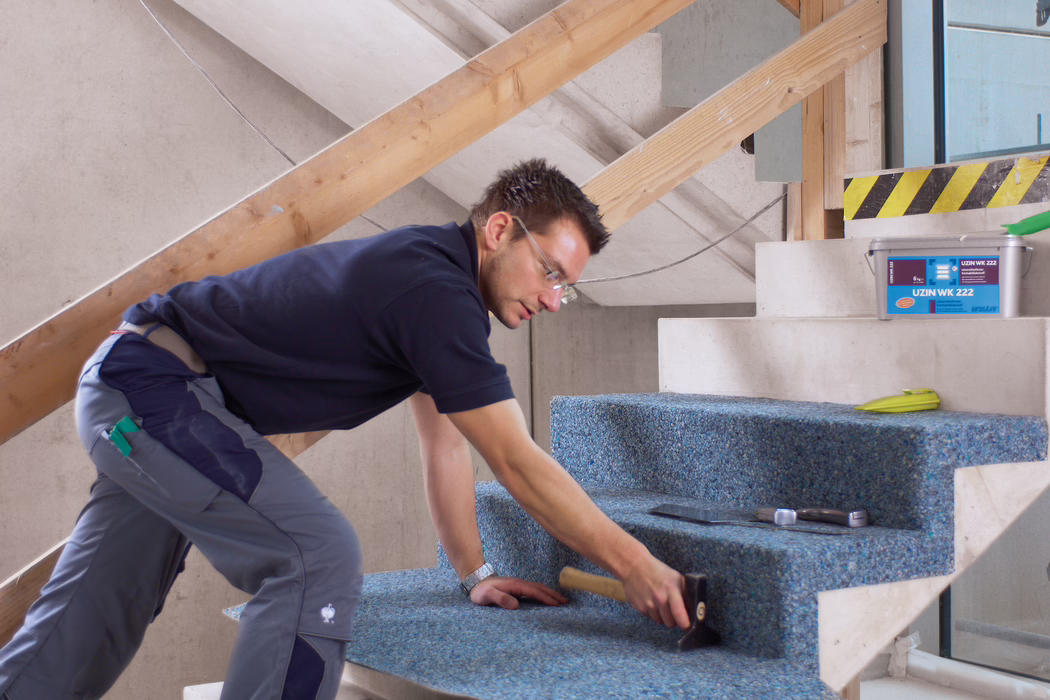 UZIN_Application_Image_WK 222_VA_4_ 2018-03 Een vakman legt een blauwe, gerecyclede vloerbedekking op betonnen traptreden in een onafgewerkt gebouwinterieur. Hij knielt en leunt naar voren, draagt een veiligheidsbril, een donkerblauw poloshirt en grijze werkbroeken, en gebruikt een rubberen hamer om het tapijt op de treden aan te drukken; in de buurt liggen een troffel, een rolmaat en een plastic bak met het opschrift 'UZIN WK 222'. De scène is goed verlicht met natuurlijk licht, tijdelijke houten leuningen zijn zichtbaar, de focus ligt op de zorgvuldige installatie en de textuur van het tapijt op de treden.
