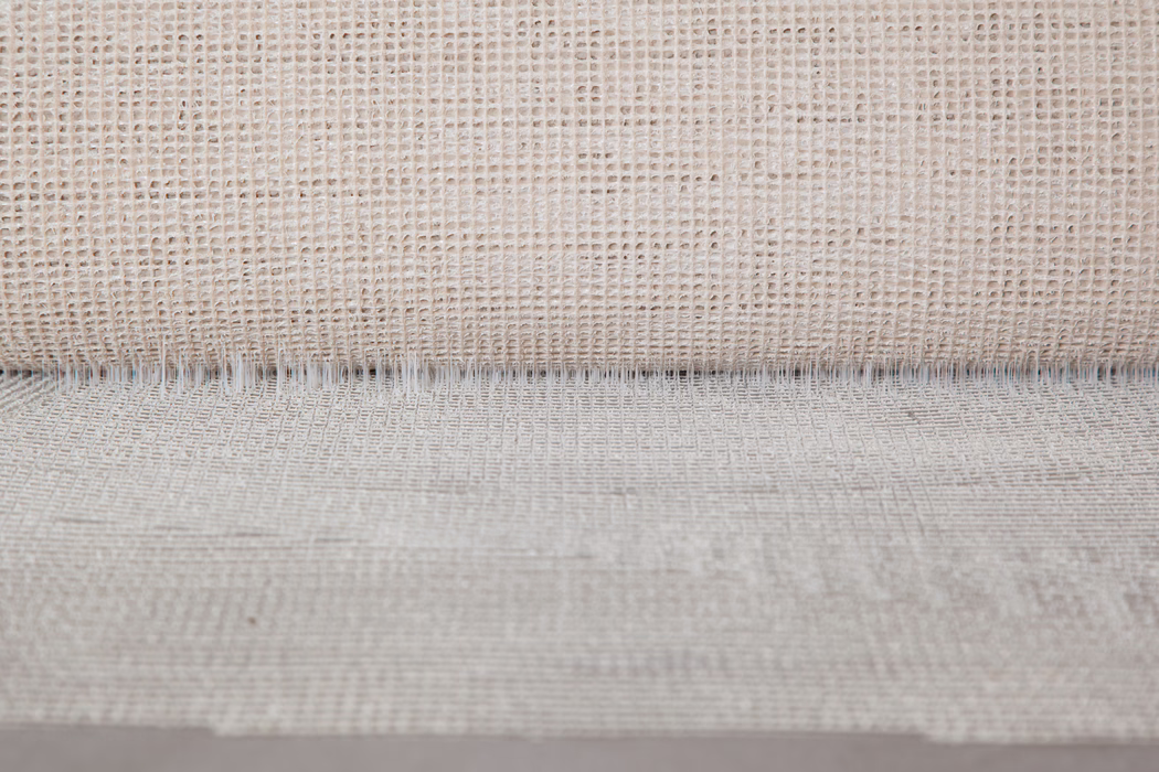 Close-up van een neutraal beige geweven stof met een open, rasterachtige structuur. Bovenaan is een opgerolde rand zichtbaar die een fijne schaduwlijn vormt; onregelmatige draden en variaties in natuurlijke vezels geven een tactiele, rustieke uitstraling. Zacht, gelijkmatig licht benadrukt de textuur en subtiele diepte.