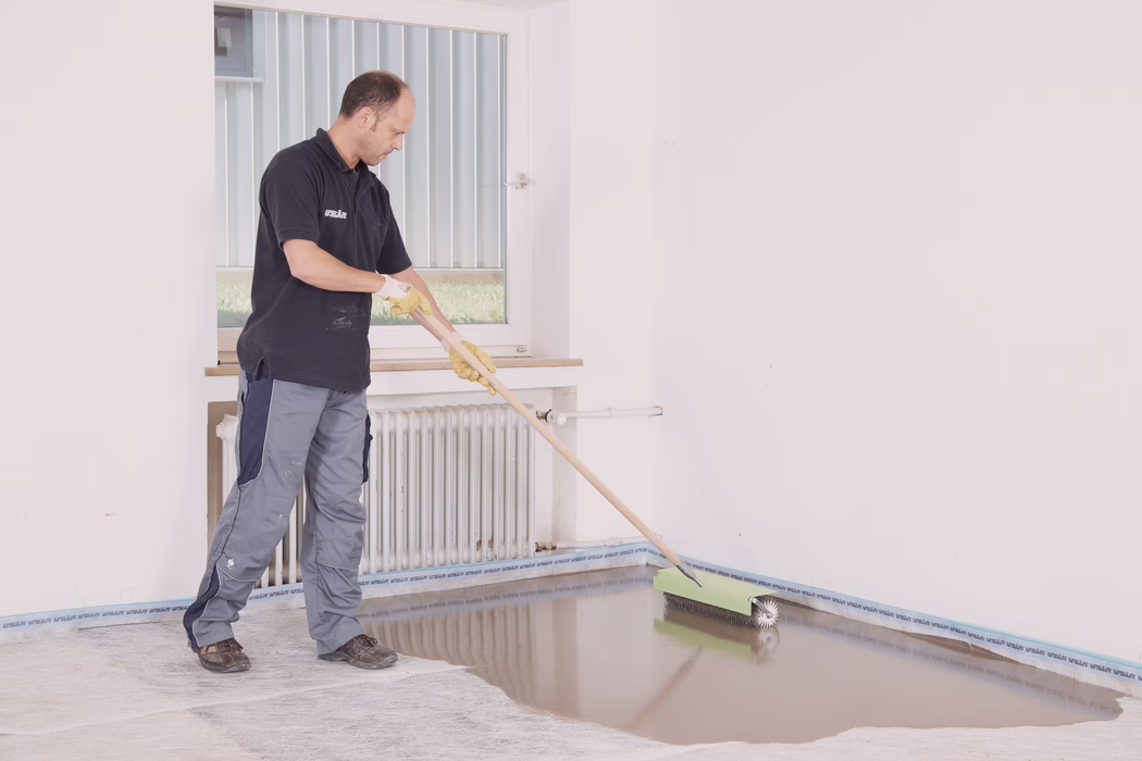 Een vakman in werkbroek, donker poloshirt en gele handschoenen brengt met een lange stalen kloprol een zelfnivellerende vloermortel aan in een lege, lichte ruimte. Hij voert de roller langs de rand van de vloer bij een witte radiator en een raam en creëert een gladde, glanzende natte laag die licht weerspiegelt. De muren zijn wit, er is afdekband langs de plint aangebracht en er zitten vlekken op de werkplek; de situatie duidt op een professionele vloerinstallatie of renovatie.
